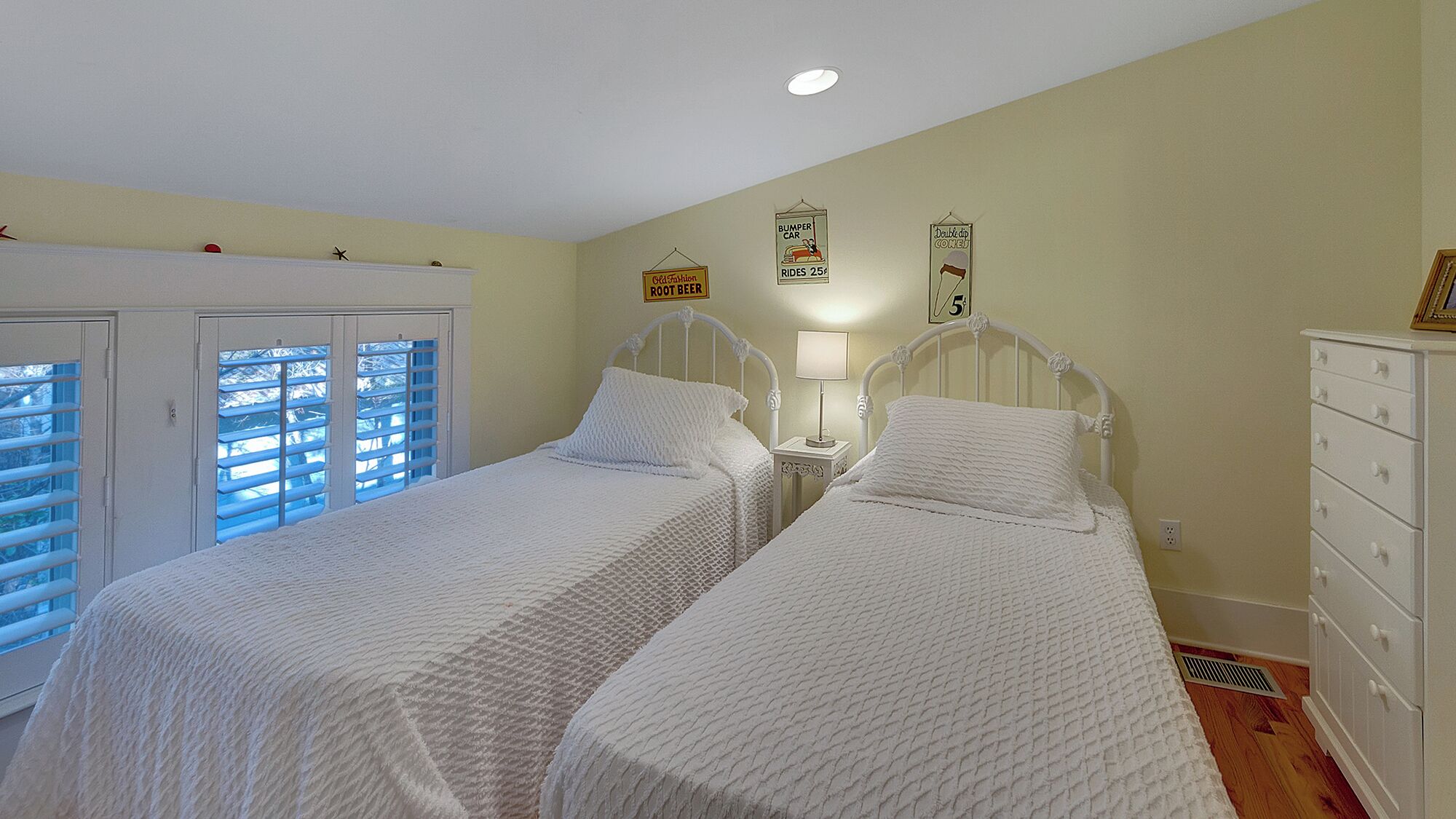 Quarto com duas camas de solteiro, mesa de cabeceira com abajur, cômoda e janela com persianas.