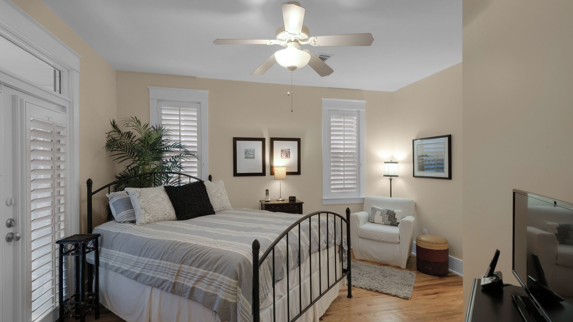 A bedroom with a bed, a ceiling fan, a nightstand, a lamp, a chair, a television, and a plant.