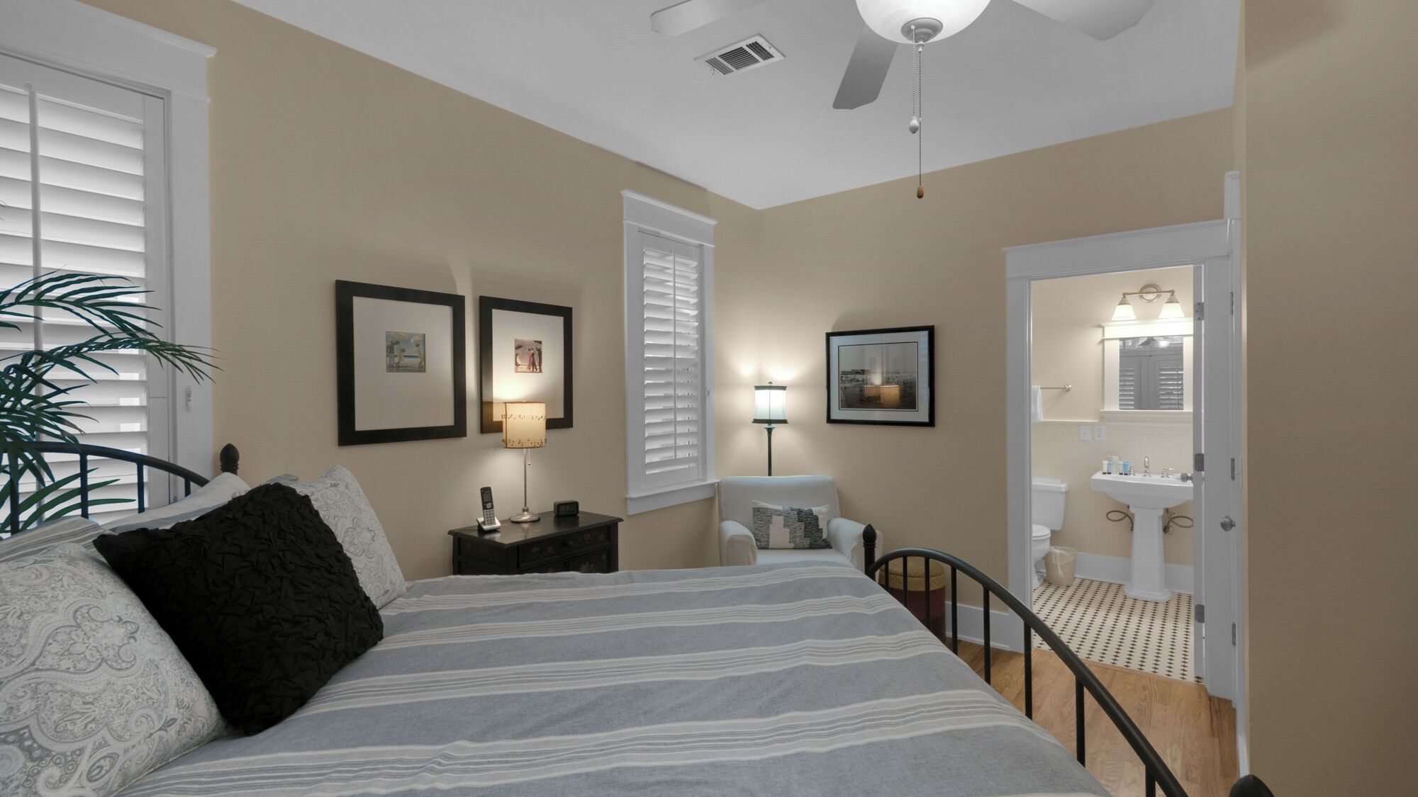 Une chambre à coucher comprenant un lit, des tables de chevet, une lampe, une chaise, une fenêtre avec des stores, un ventilateur de plafond, et une salle de bain visible à travers une porte ouverte.