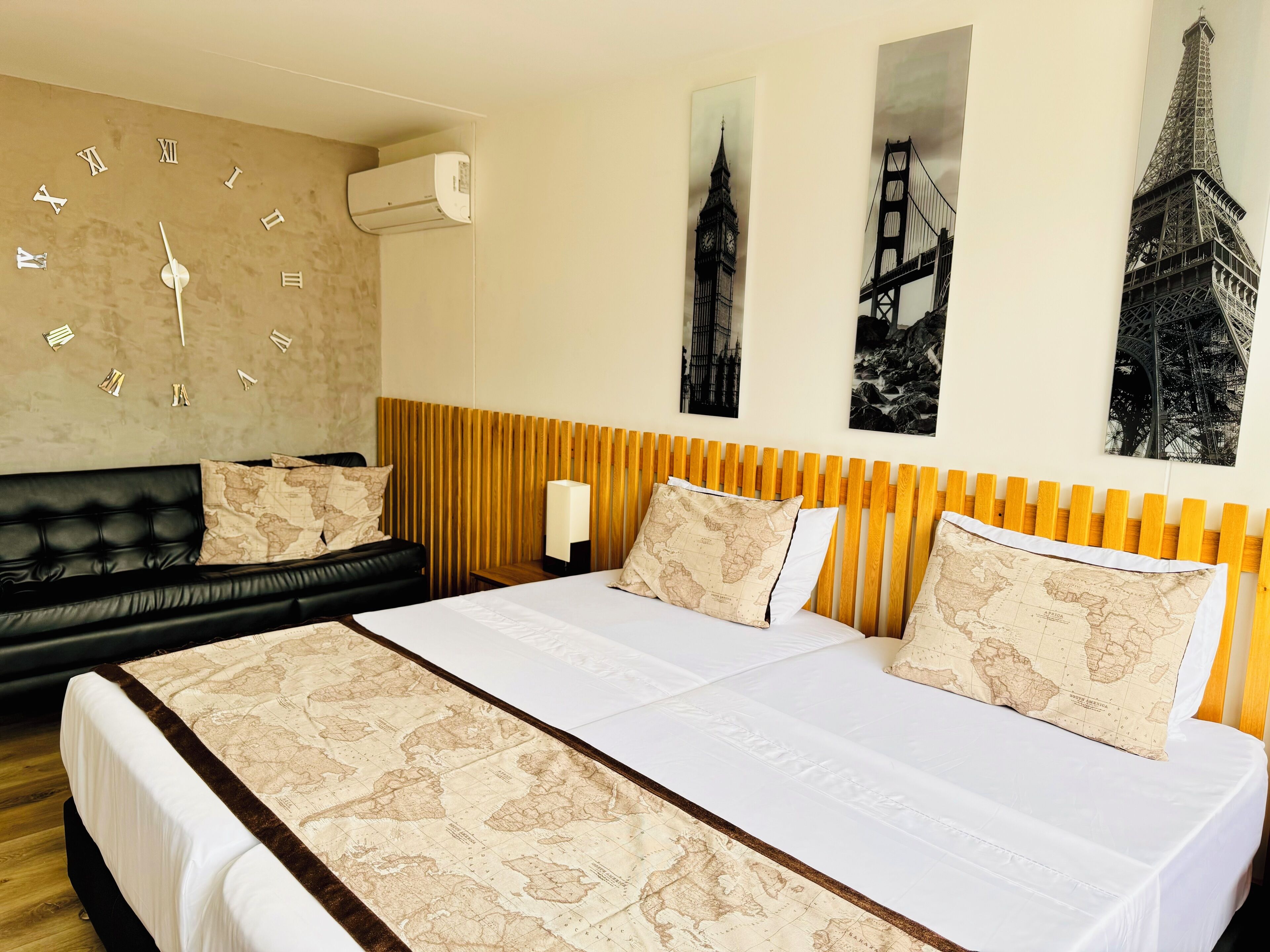 Habitación de hotel con una cama grande, un sofá de cuero negro, cabecera de madera y obras de arte en la pared que representan monumentos famosos.