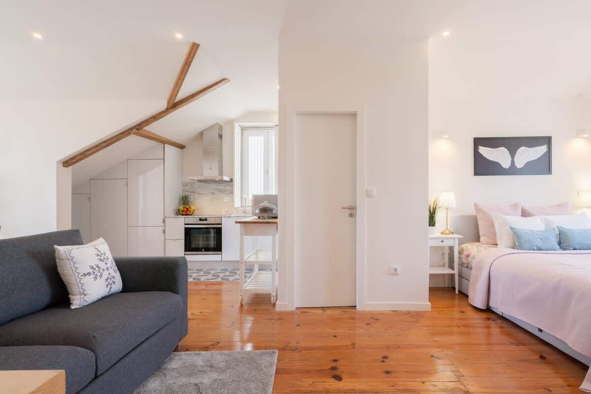 A modern apartment with a grey sofa, a bed with a pink and blue duvet, and a kitchen in the background.
