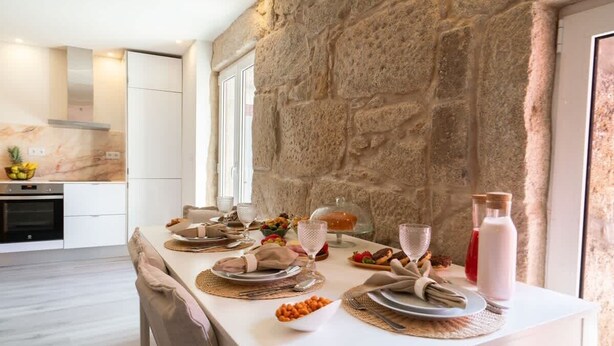 Uma sala de jantar com parede de pedra, cozinha ao fundo e mesa posta para refeição.