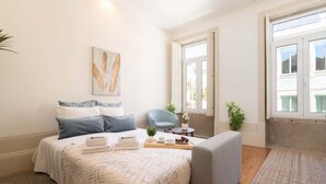 Quarto bem organizado com cama, sofá cinzento, pequena mesa com bandeja e uma cadeira.