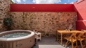 Um muro de pedra, um vigem vermelho, uma banheira de hidromassagem, uma mesa com cadeiras e uma pequena mesa com uma garrafa e uma planta.
