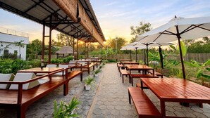 Un patio al aire libre con mesas y sillas de madera, una zona de estar cubierta y plantas en macetas.
