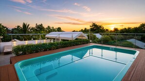 Una piscina rectangular con cubierta de madera, rodeada por una barandilla de vidrio, y una estructura tipo marquesina blanca al fondo.