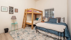 A bedroom with a bunk bed, a single bed, a small table with a lamp, a trash bin, and a window with blinds. - Wide open spaces getaway (Concho)