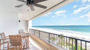 Ein Balkon mit Deckenventilator, Holzstühlen und einem Tisch. Blick auf einen Strand mit weißem Sand und klarem, blauem Wasser.