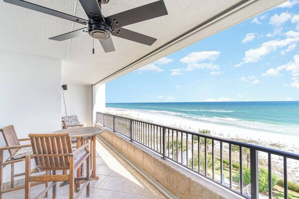 A balcony with a ceiling fan, wooden chairs, and a table. It overlooks a beach with white sand and clear blue water.