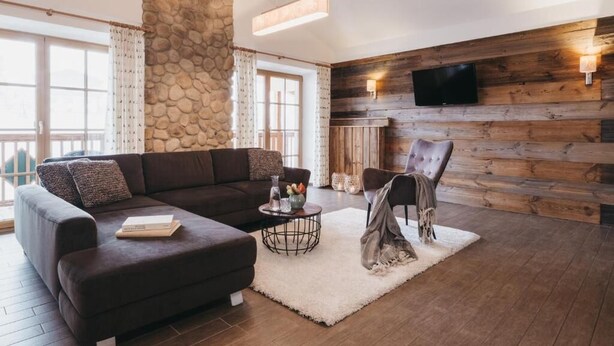 Un salon chaleureux avec un mur en pierre, un lambris en bois, un canapé, un fauteuil, une table basse et une cheminée.