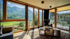 Une pièce spacieuse avec de grandes fenêtres donnant sur les montagnes, un coin salon confortable avec un fauteuil et une petite table, et un plafond en bois.