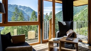 Une chambre moderne dotée de grandes fenêtres offrant une vue sur les montagnes, d’un balcon avec une balustrade et d’un coin salon confortable avec un canapé et un fauteuil.