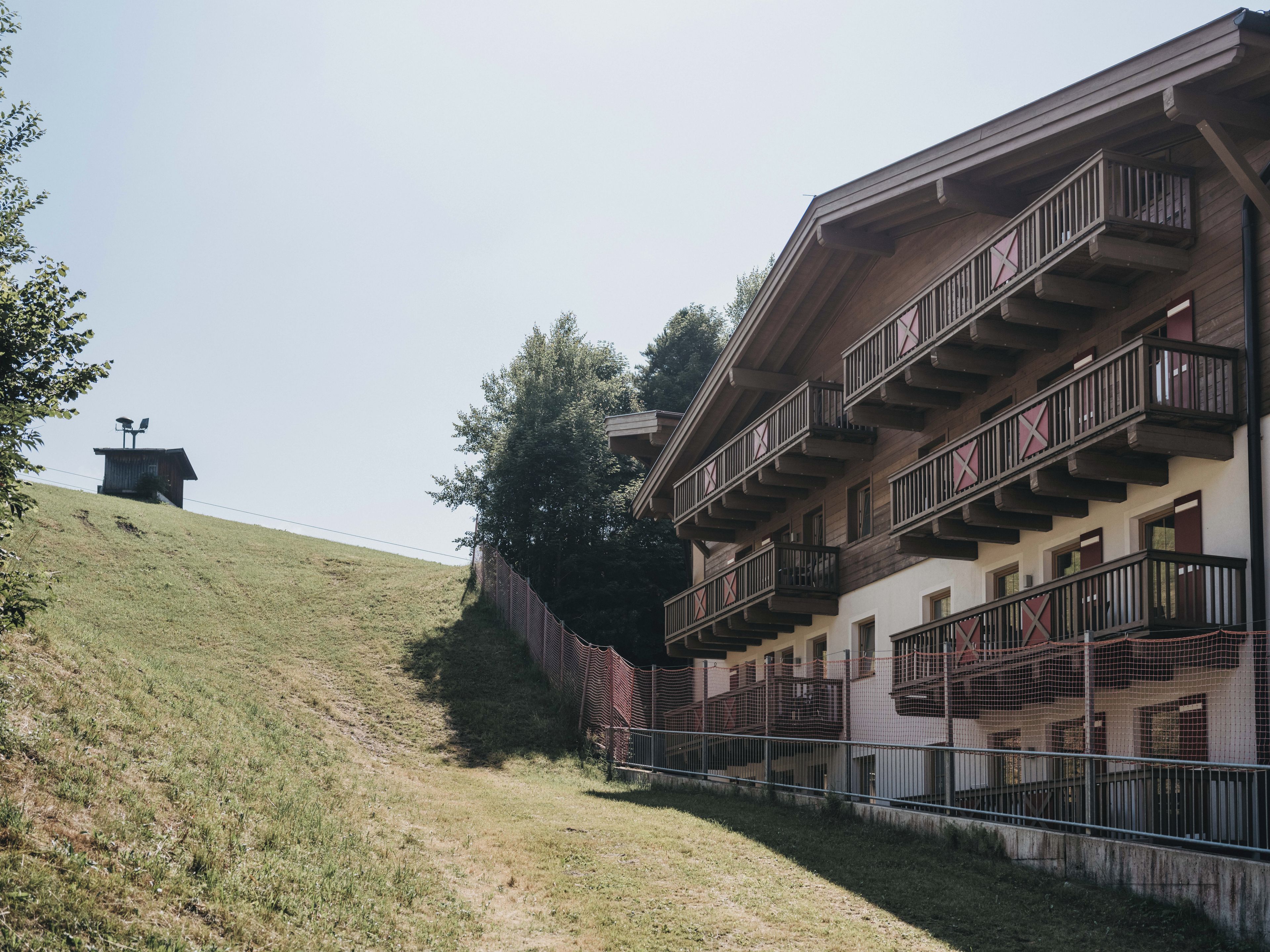 A multi-story building with balconies, a grassy hill, and a small structure in the background.