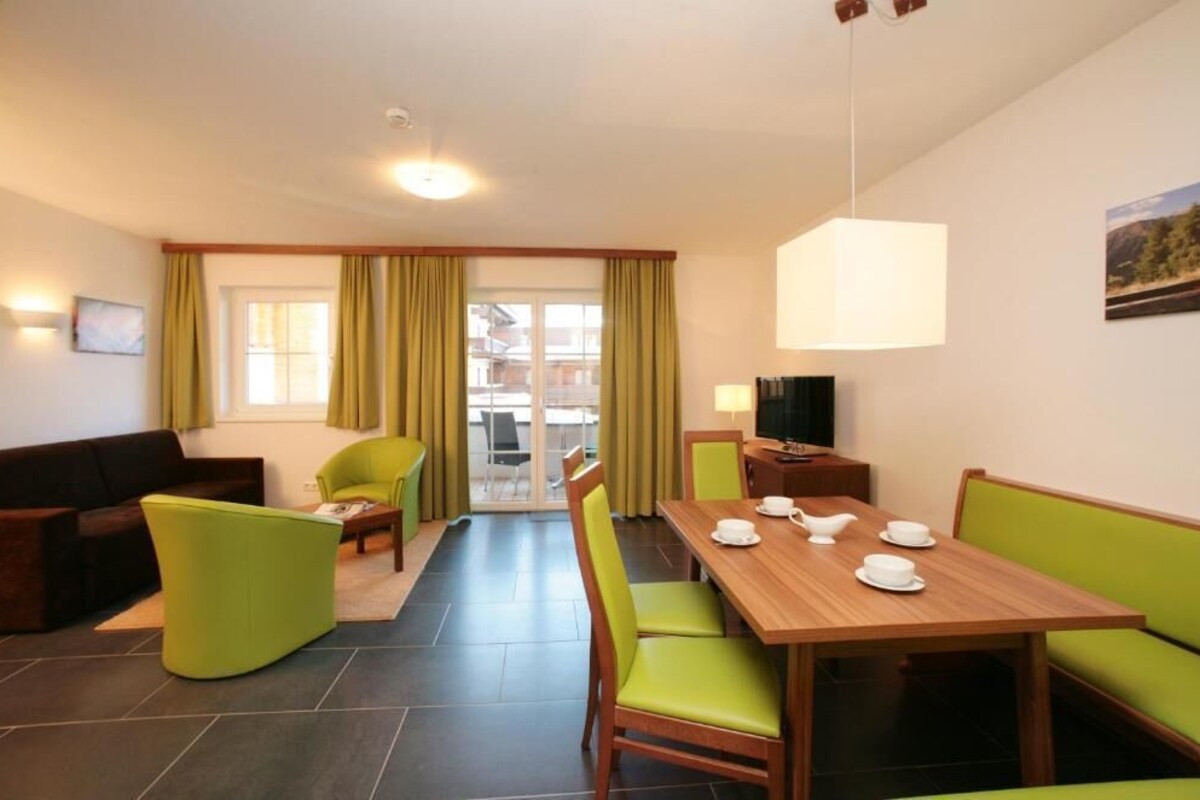 Een moderne woonkamer met een houten eettafel voor vier personen, groene stoelen, een bank en een televisie.