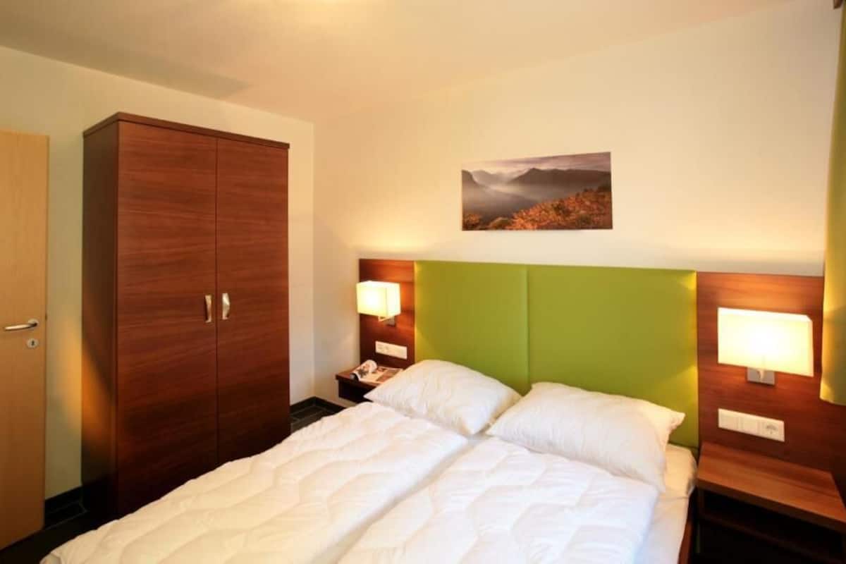 Une chambre d’hôtel comprenant un lit, deux tables de chevet, une petite table, une armoire et une lampe fixée au mur.