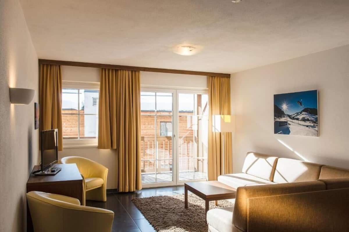 Une chambre d’hôtel avec un canapé, un fauteuil et un bureau équipé d’un ordinateur. Il y a une grande fenêtre avec des rideaux, un tableau au mur et une vue sur un bâtiment à l’extérieur.
