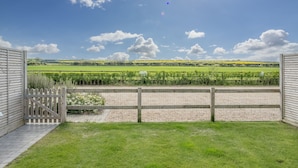 A view from a balcony with a wooden fence, a paved pathway, and a field of crops in the background. - Kingfishers Cove, Titchwell, Norfolk (Wells-next-the-Sea)