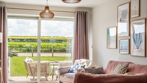 A modern living room with a sofa, a coffee table, and a wicker chair. There is a large window with curtains, and artwork on the wall. - Kingfishers Cove, Titchwell, Norfolk (Wells-next-the-Sea)