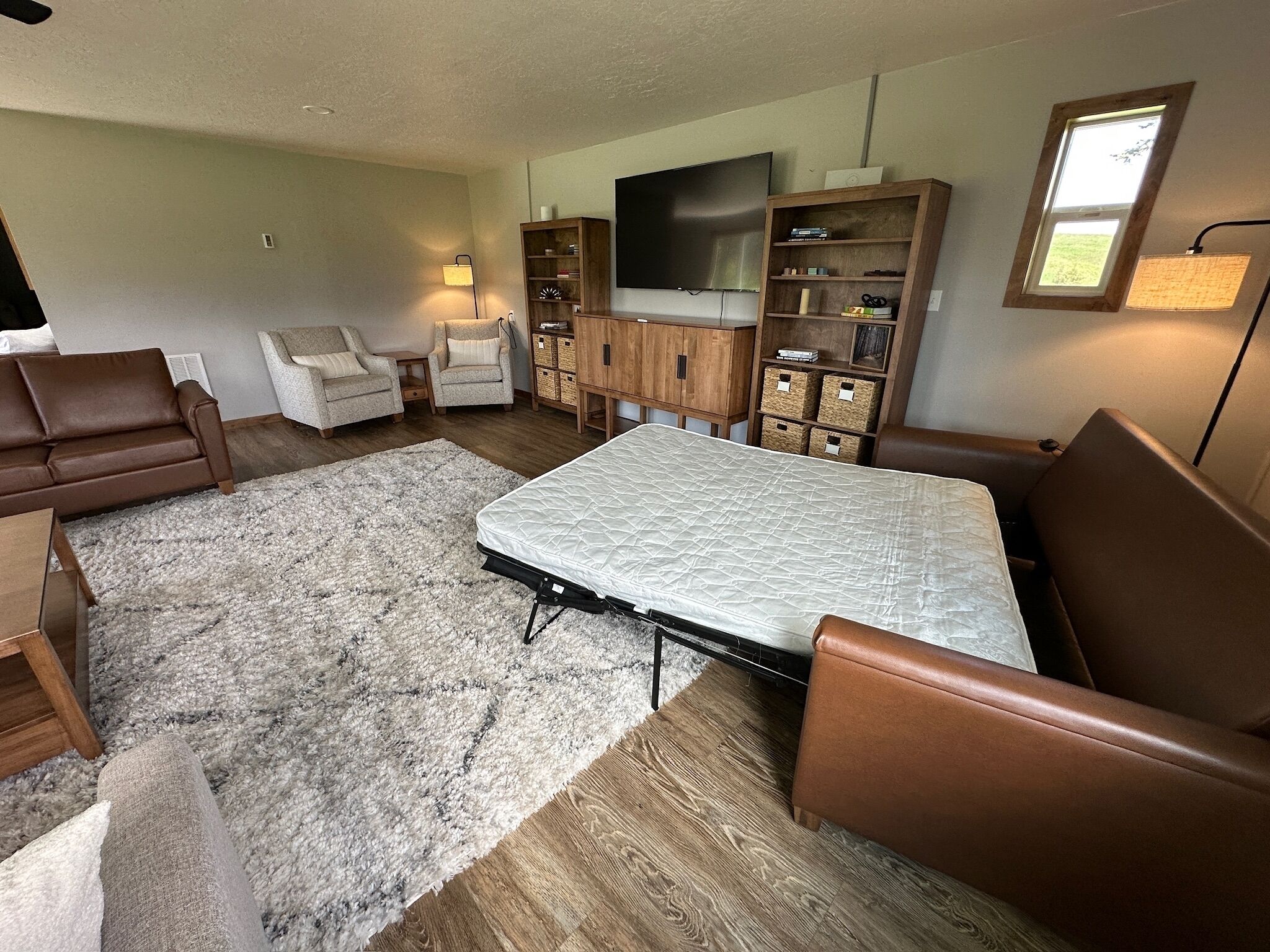 A living room with a sofa, armchairs, a TV, wooden cabinets, and a large window.