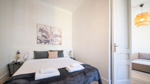 Una cama bien hecha con cubierta negra, sábanas blancas y dos toallas dobladas. Hay dos apliques de luz en la pared y una puerta que da a otra habitación.
