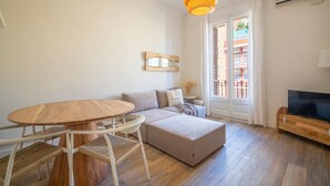 Una sala de estar moderna con una mesa redonda de madera, sillas de mimbre blanco, un sofá beige y un televisor sobre un mueble de madera.
