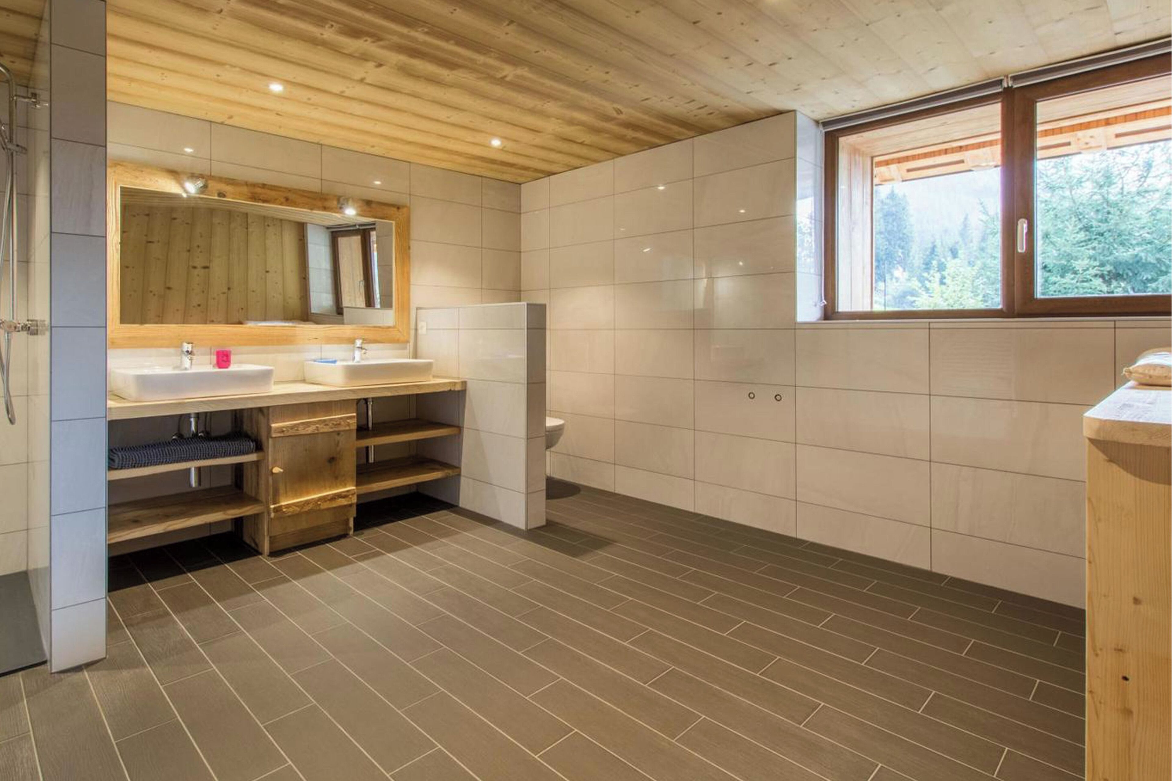 Un baño moderno con techo de madera, dos lavabos, un espejo grande y una ventana con vista a árboles.