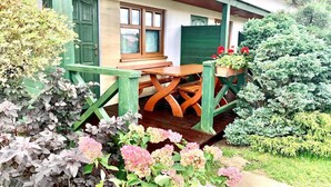 Eine hölzerne Bank unter einer überdachten Terrasse mit einer grünen Tür und Fenstern, umgeben von verschiedenen Pflanzen und Blumen.