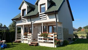 Een tweelaags huis met een balkon, een veranda met houten bankjes en een schommel in de tuin.