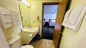 Une petite salle de bain avec des toilettes, un lavabo et un miroir. Des serviettes sont accrochées au mur, et une porte donne sur une chambre.