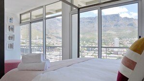 Habitación de hotel con una cama grande, vistas a un paisaje urbano y montañas, y una ventana con marco de metal.
