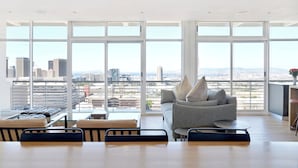 Amplia habitación con un ventanal que ofrece vistas a la ciudad, un sofá gris, una pequeña mesa redonda y un rincón de estar con sillas.