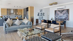 Una sala moderna con un sofá gris, una mesa de centro de vidrio y un reposapiés de cuero. Hay un comedor con una mesa y sillas de madera.