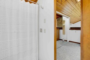 Un pasillo estrecho con techo de madera y pared blanca. Una cortina blanca cuelga a la izquierda, y una puerta da paso a una habitación con pared de piedra y estante de madera.
