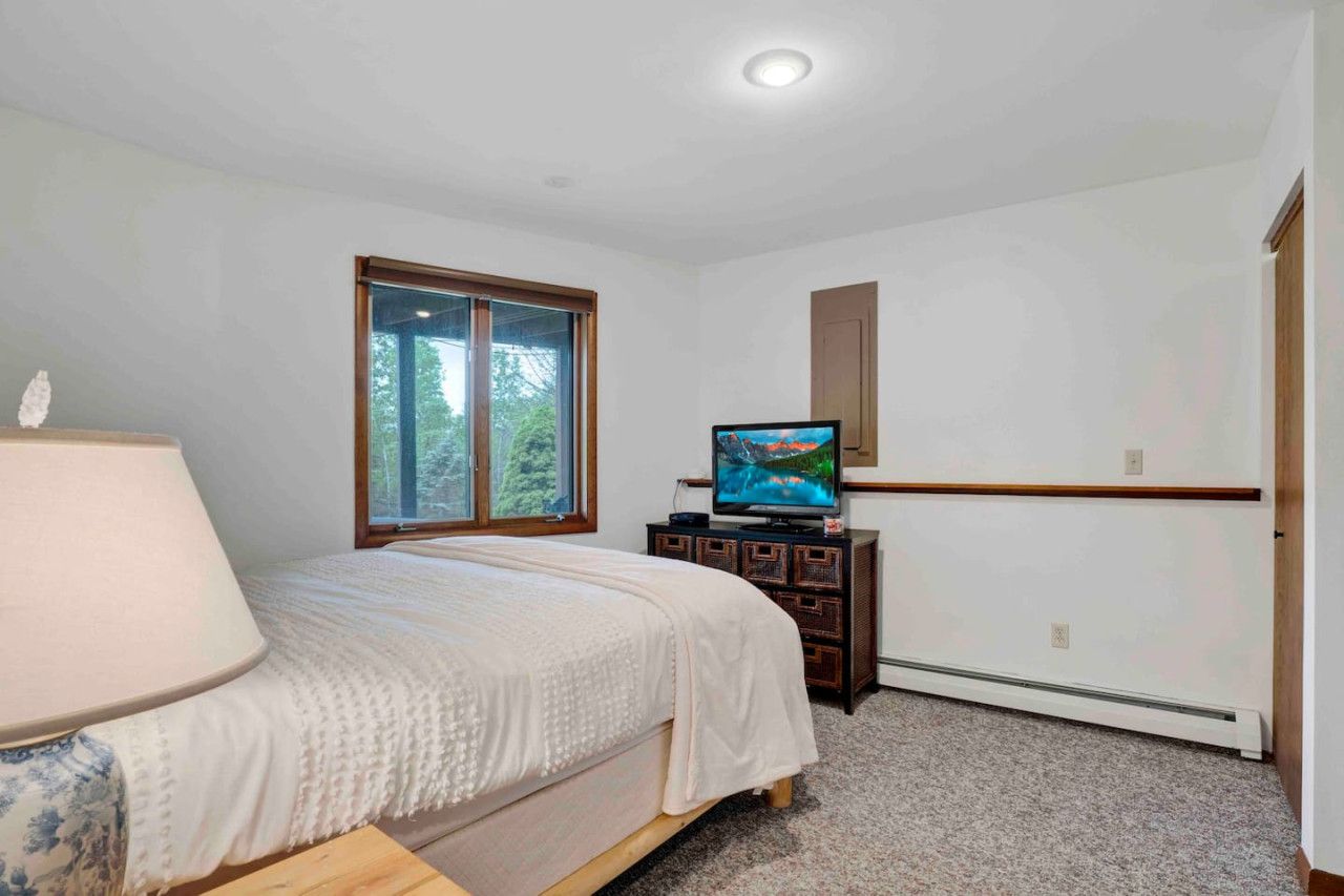 A bedroom with a bed, a television on a wooden cabinet, a window with a view of trees, and a ceiling light.