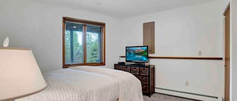 A bedroom with a bed, a television on a wooden cabinet, a window with a view of trees, and a ceiling light.