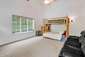A room with a bunk bed, a small table, a black couch, and a window with a view of greenery.