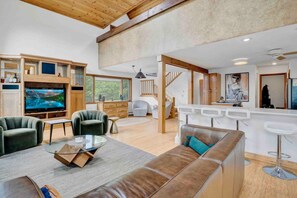 A spacious living area with a large flat-screen TV, comfortable seating, a kitchen area, and a staircase leading to another level.