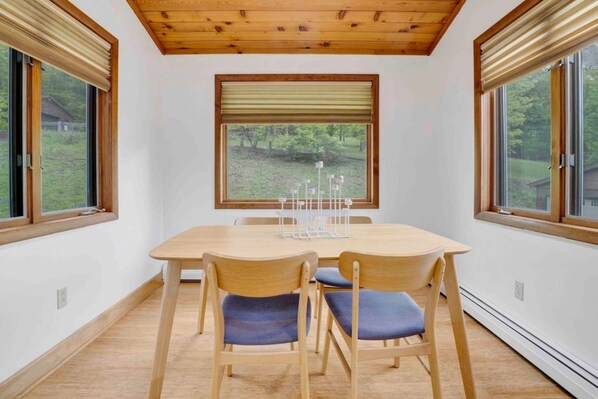 A dining area with a wooden table and chairs, large windows with blinds, and a view of greenery outside.