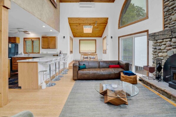 Un amplio espacio habitable con chimenea, una cocina con barra y un comedor con techo alto y luz natural.