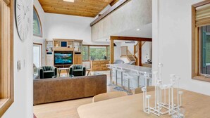 A modern living room with a large TV, comfortable seating, a kitchen area, and a dining space with bar stools.