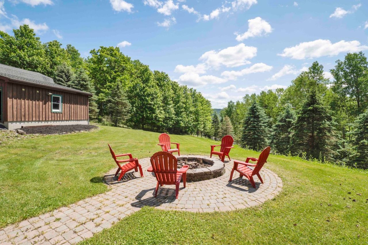 A circular fire pit area with red Adirondack chairs and a brick pathway leading to a wooden building.