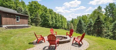 A circular fire pit area with red Adirondack chairs and a brick pathway leading to a wooden building.