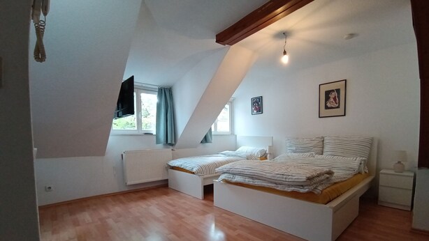 Ein Schlafzimmer mit schrägen Wänden, einem Bett mit gestreifter Bettwäsche, einem Nachttisch, einem Fenster mit Vorhängen und einer Heizung.
