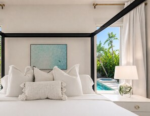 A bedroom with a bed, a nightstand, a lamp, a painting, and a view of a pool.