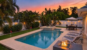 A swimming pool with a hot tub, surrounded by palm trees and a patio area with chairs and a table.