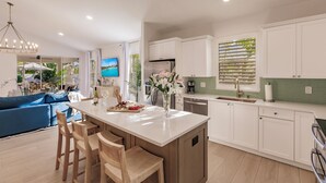 A modern kitchen with white cabinets, a central island with bar stools, a dining table, and a view of the outdoors through large windows. - LUXE, Beach, Hot Tub, Putt Putt, 2 Mstr Suites, 3 Kings, Hot Tub, Bunkbds, Pets (Naples)