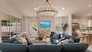 A modern living room with a large sofa, a coffee table, and a chandelier. There is a television mounted on the wall and a kitchen area visible in the background. - LUXE, Beach, Hot Tub, Putt Putt, 2 Mstr Suites, 3 Kings, Hot Tub, Bunkbds, Pets (Naples)