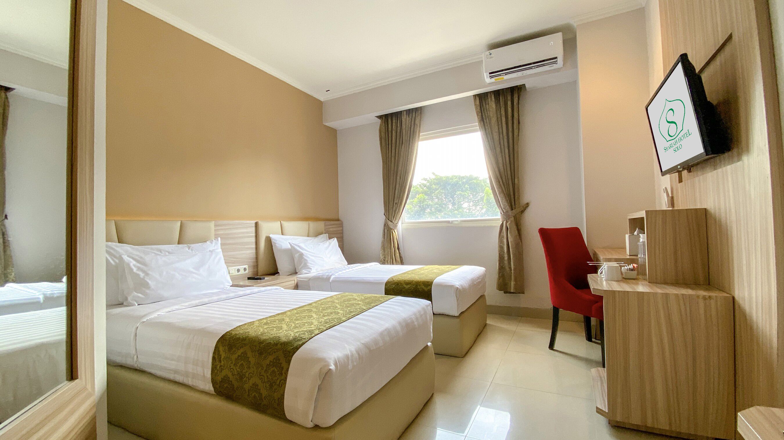 A hotel room with two beds, a desk, a chair, an air conditioning unit, and a TV mounted on the wall.