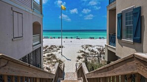 Une vue sur une plage, prise depuis un bâtiment surmonté d’un mât, avec un escalier en bois et une plage de sable parsemée de parasols.