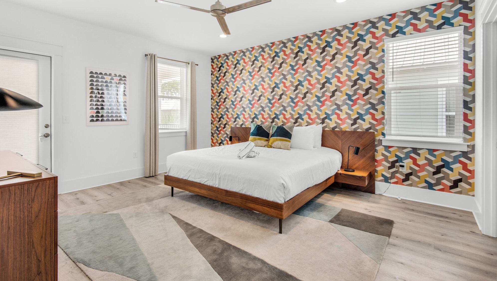 A modern bedroom with a wooden bed, a ceiling fan, a patterned wall, and a window with blinds.